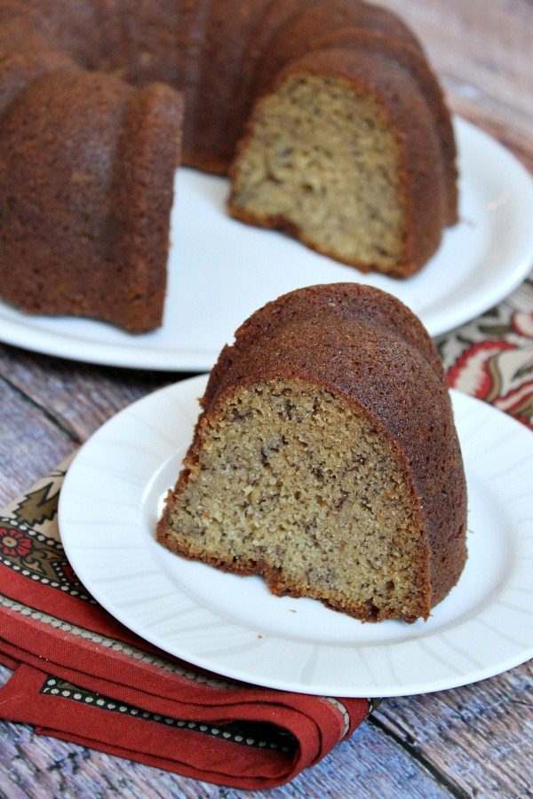 classic banana bundt cake on a white cake platter with a slice of cake on a white plate sitting in front of it. patterned rust colored cloth napkin under the plate