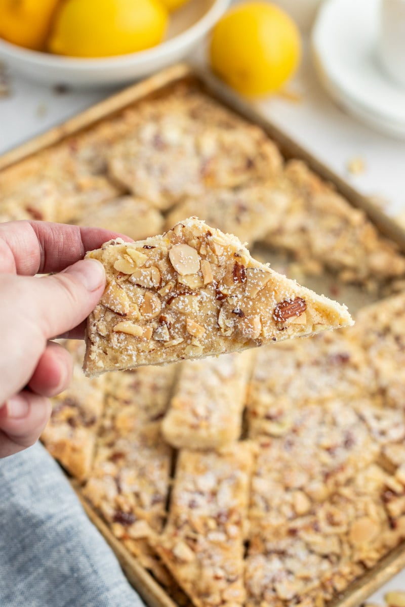 hand holding a piece of lemon almond cookie brittle over the pan of the rest of the brittle