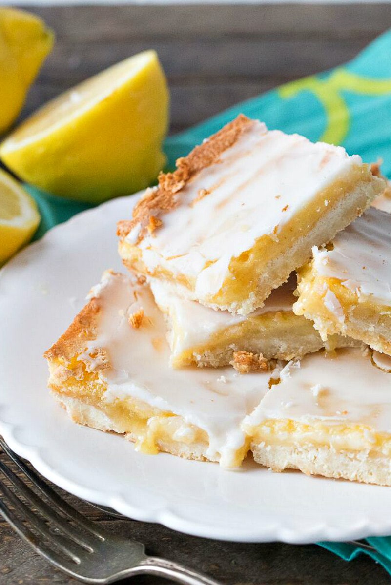 Best Lemon Bars stacked on a white plate. Fork in front. Fresh lemons and a turquoise and green patterned napkin in the background