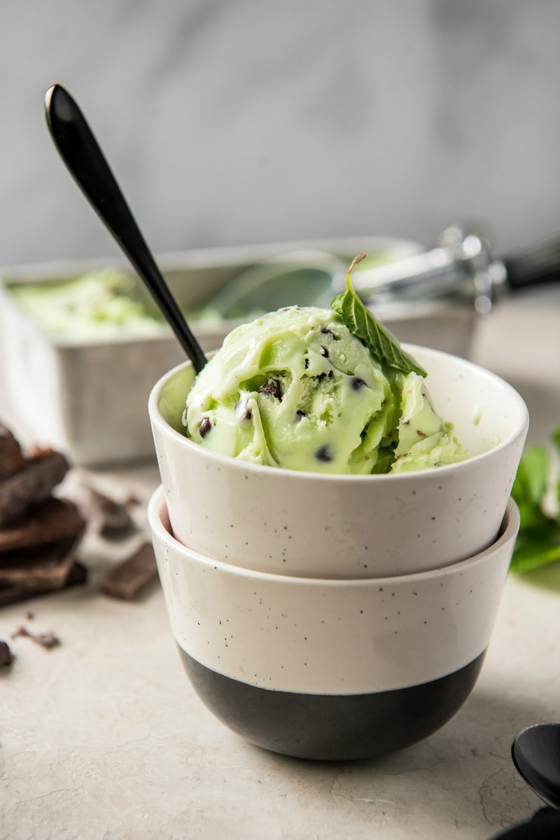 dish of mint chocolate chip ice cream with a spoon with chocolate and the ice cream container in the background