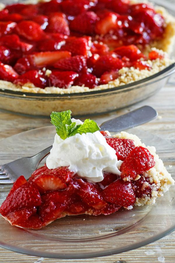 Slice of Fresh Strawberry Pie displayed on a glass plate with whipped cream and fresh mint on top. Fork on the plate. Whole strawberry pie in the background.