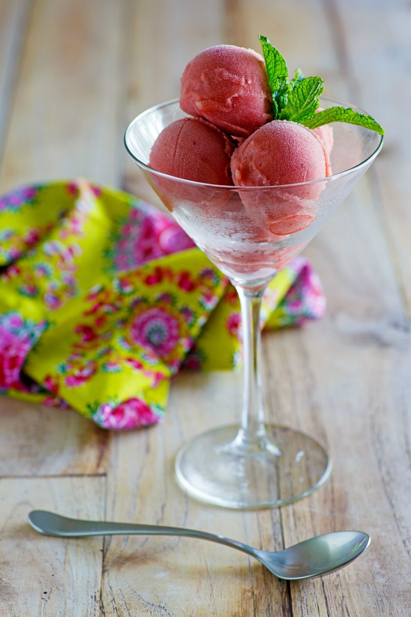 Scoops of Watermelon Sorbet in a martini glass garnished with fresh mint. spoon laying on table in front of glass. patterned pink and green cloth napkin displayed in background