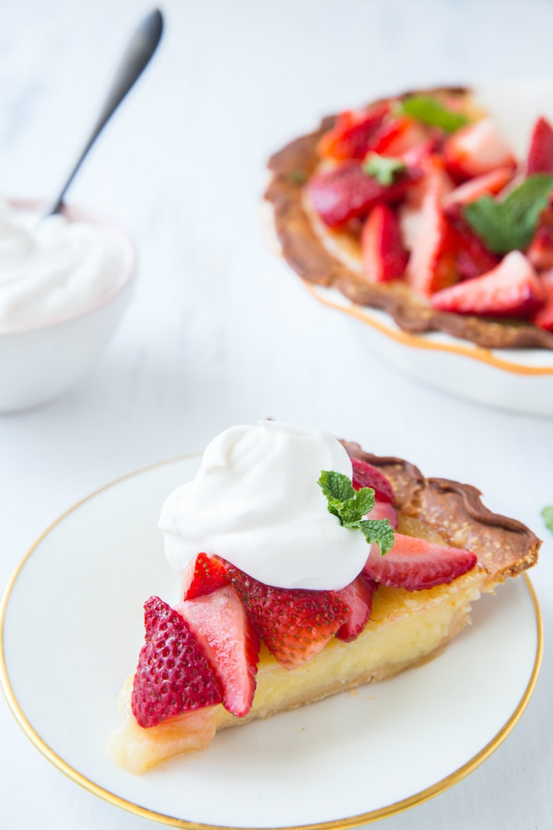 slice of strawberry lemon buttermilk pie on a white plate, garnished with whipped cream. The rest of the pie is in the background