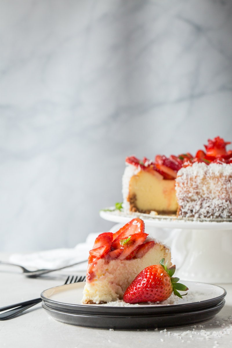 slice of strawberry coconut cheesecake on a white plate with whole cheesecake on display in the background