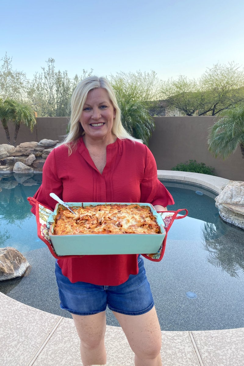 RecipeGirl holding a pan of Baked Penne with Italian Sausage standing in front of a pool in backyard