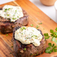 two steaks on a cutting board topped with blue cheese sauce
