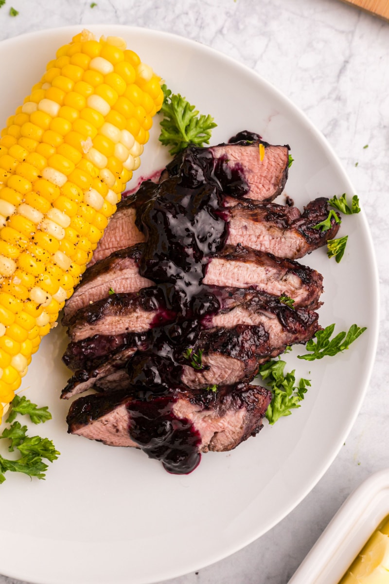 sliced pork tenderloin on a plate with corn topped with blueberry bbq sauce