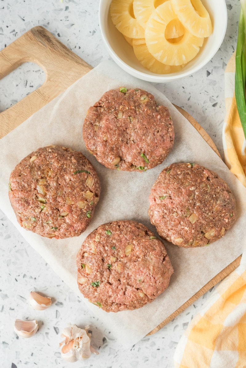 Teriyaki Burgers recipe from RecipeGirl.com #teriyaki #burger #burgers #recipe #RecipeGirl four teriyaki burger patties on a cutting board with sliced pineapple on the side