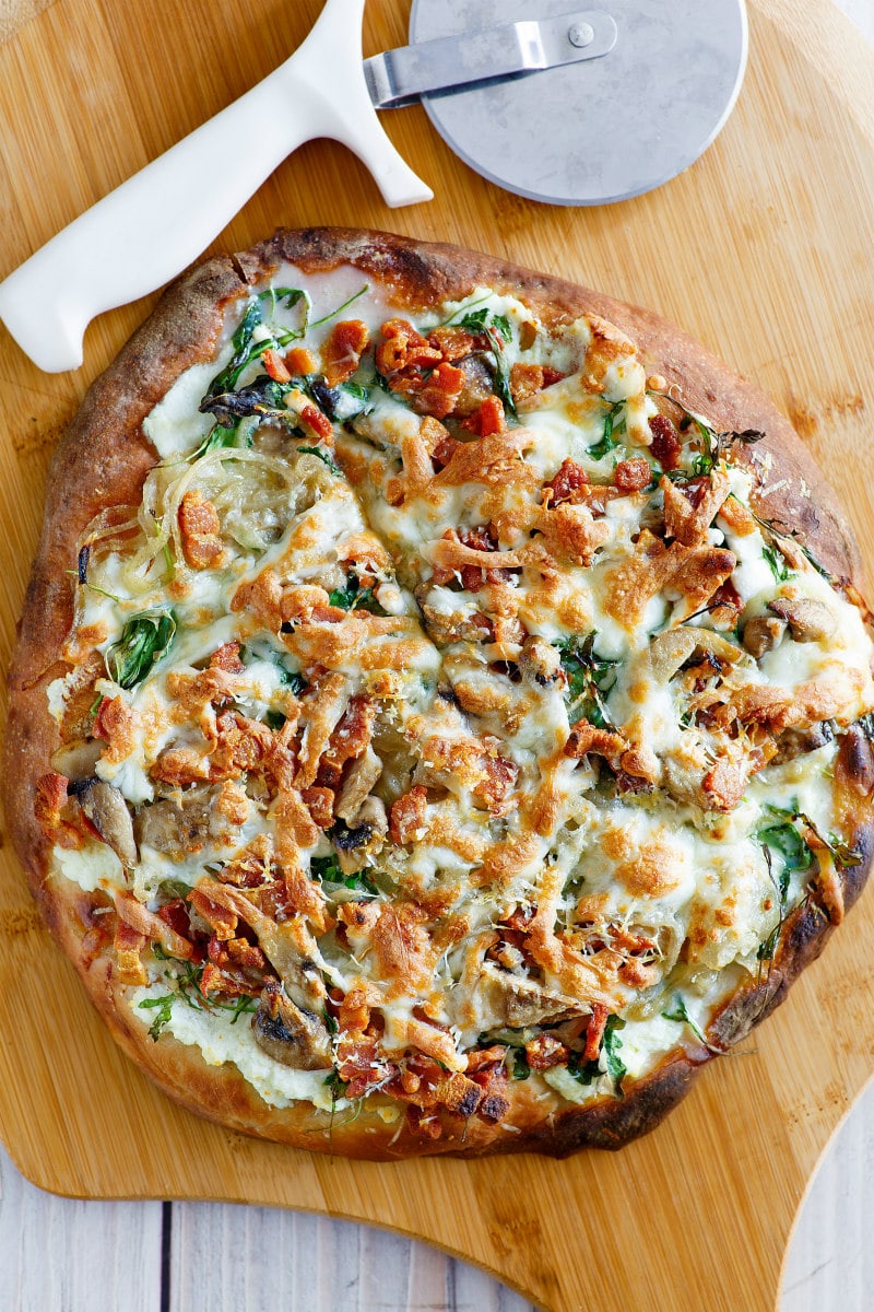 overhead shot of white pizza with arugula, bacon and mushrooms on a cutting board with a pizza cutter on the side