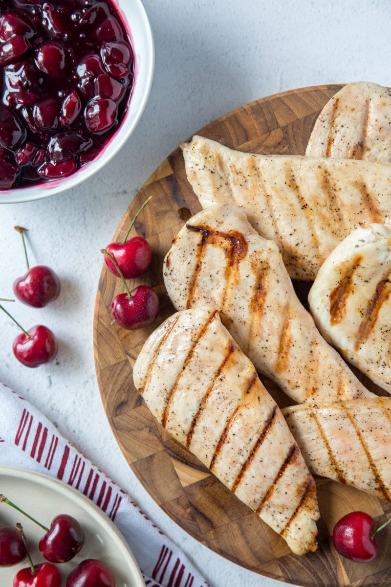 grilled chicken breasts on a wooden board. a bowl of cherry sauce in the background with some fresh cherries scattered around