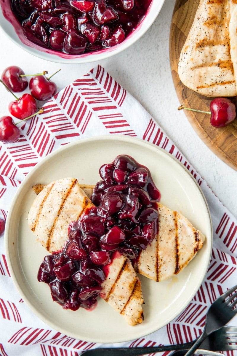 overhead shot of a white plate on a patterned red and white napkin with grilled chicken and cherry sauce on top. bowl of cherries and fresh cherries and more grilled chicken in the background