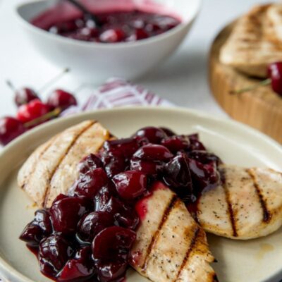 grilled chicken on a white plate with cherry sauce . bowl of cherry sauce in the background