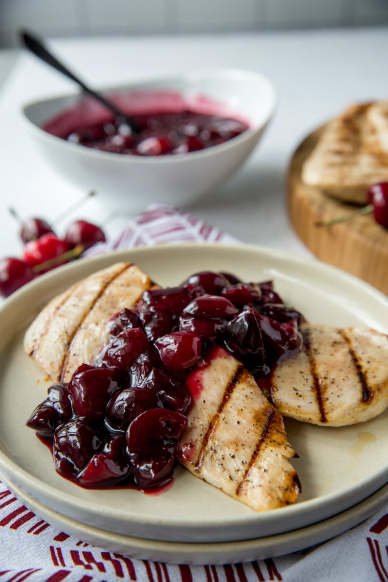 grilled chicken on a white plate with cherry sauce . bowl of cherry sauce in the background