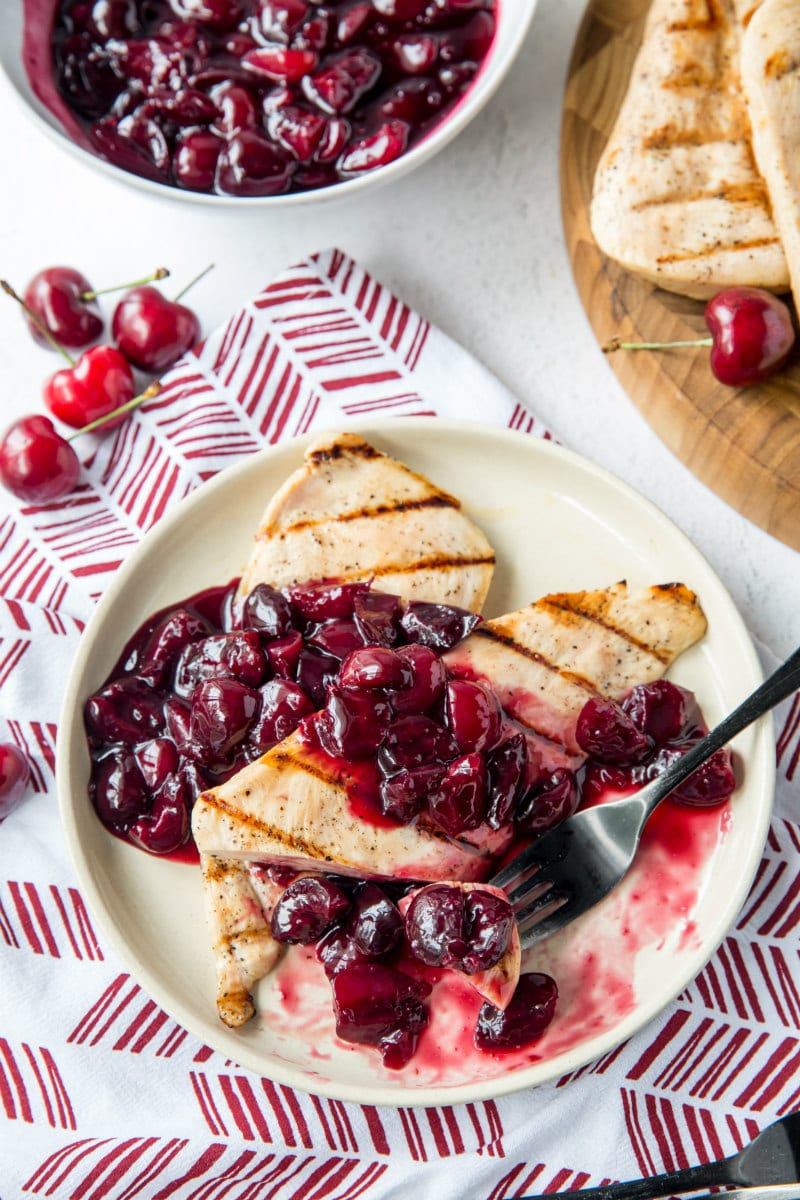 overhead shot of a serving of grilled chicken on a white plate served with cherry sauce. Fork on plate and red/white patterned napkin underneath