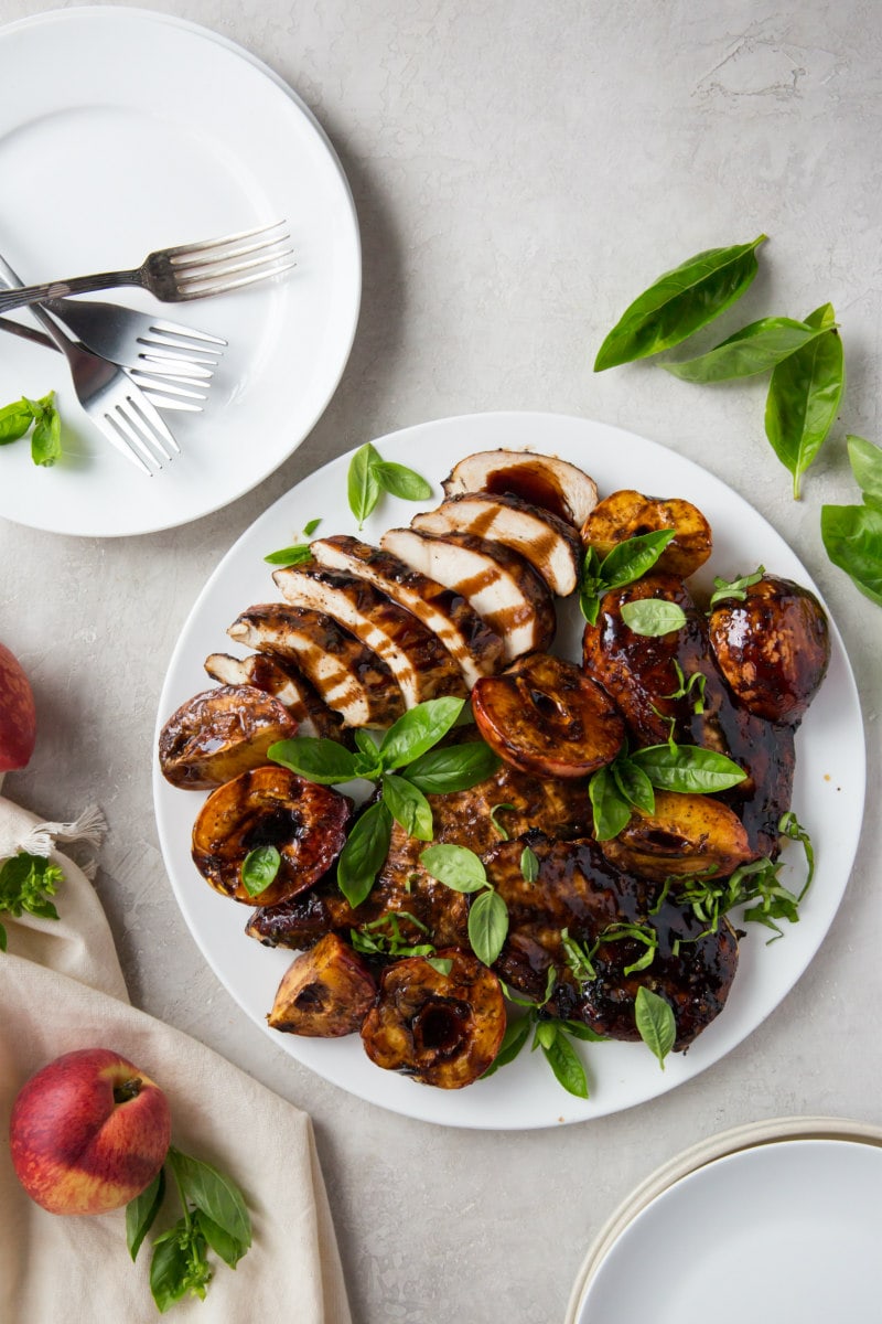 grilled chicken and peaches on a white plate with a plate of forks on the side and garnished with basil