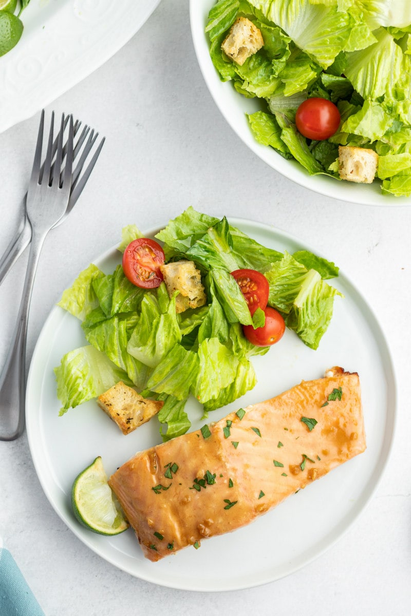 overhead shot of white dinner plate with salmon and salad. forks on the side and plate of salad on the side.