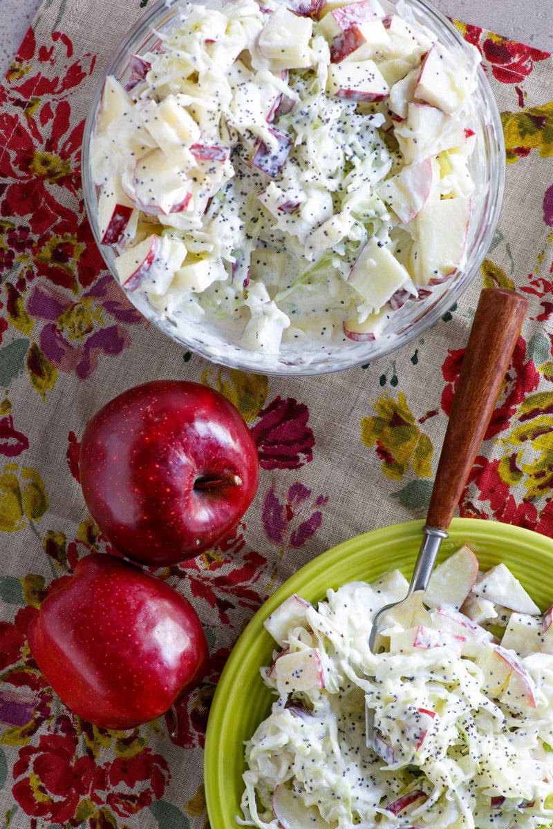 overhead shot of apple cole slaw in a serving bowl and on a green salad plate. red apples on the side. set on a floral tablecloth