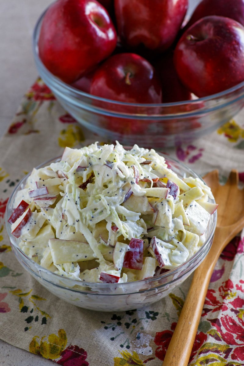 apple cole slaw in a bowl and apples in another bowl. wooden spoon on the side. set on a floral napkin