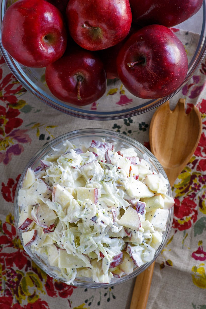 overhead shot of apple cole slaw in a bowl with red apples in another bowl. wooden spoon on the side. set on a floral patterned napkin