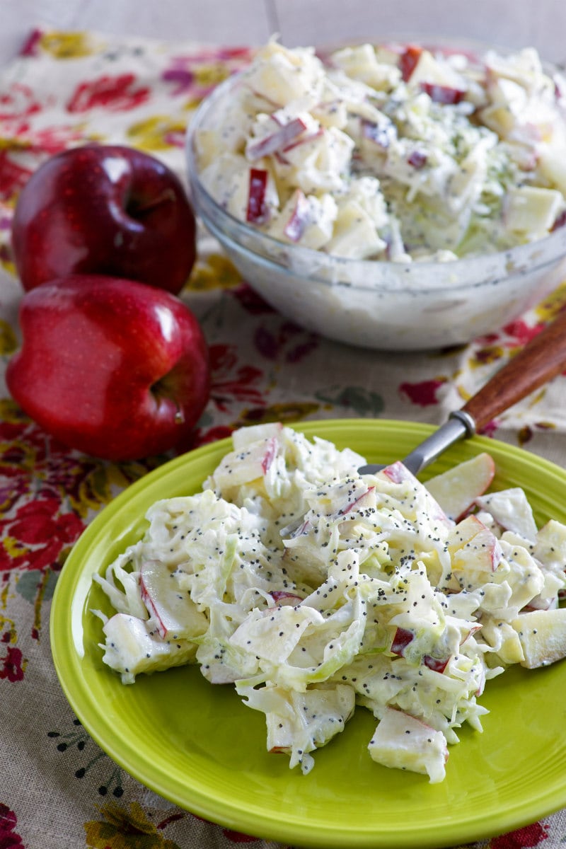 serving of apple cole slaw on a green plate. cole slaw bowl in background as well as red apples