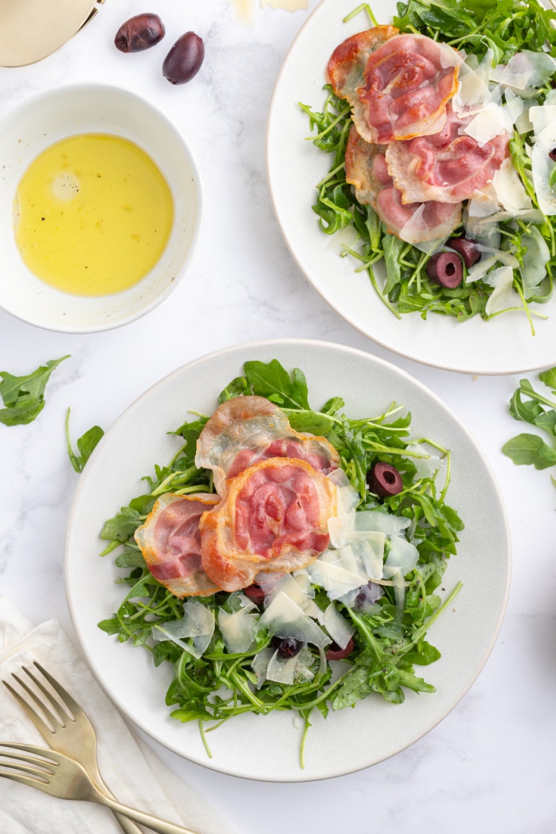 two plates of arugula salad with olives pancetta and parmesan