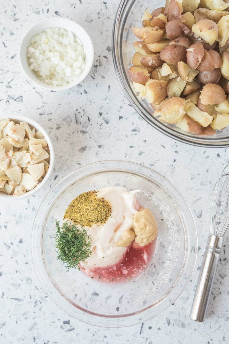 overhead shot of ingredients in bowls for california potato salad. dressing ingredients set into a glass bowl, plus bowl of artichoke hearts, and onion and potatoes on display