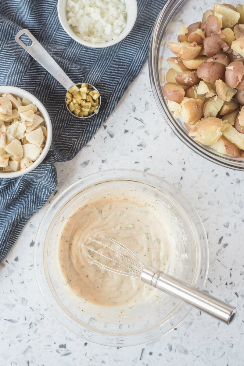 overhead shot of ingredients for california potato salad with a bowl of dressing and bowl of potatoes, as well as a bowl of artichoke hearts, dill pickle and onion, set on a blue napkin