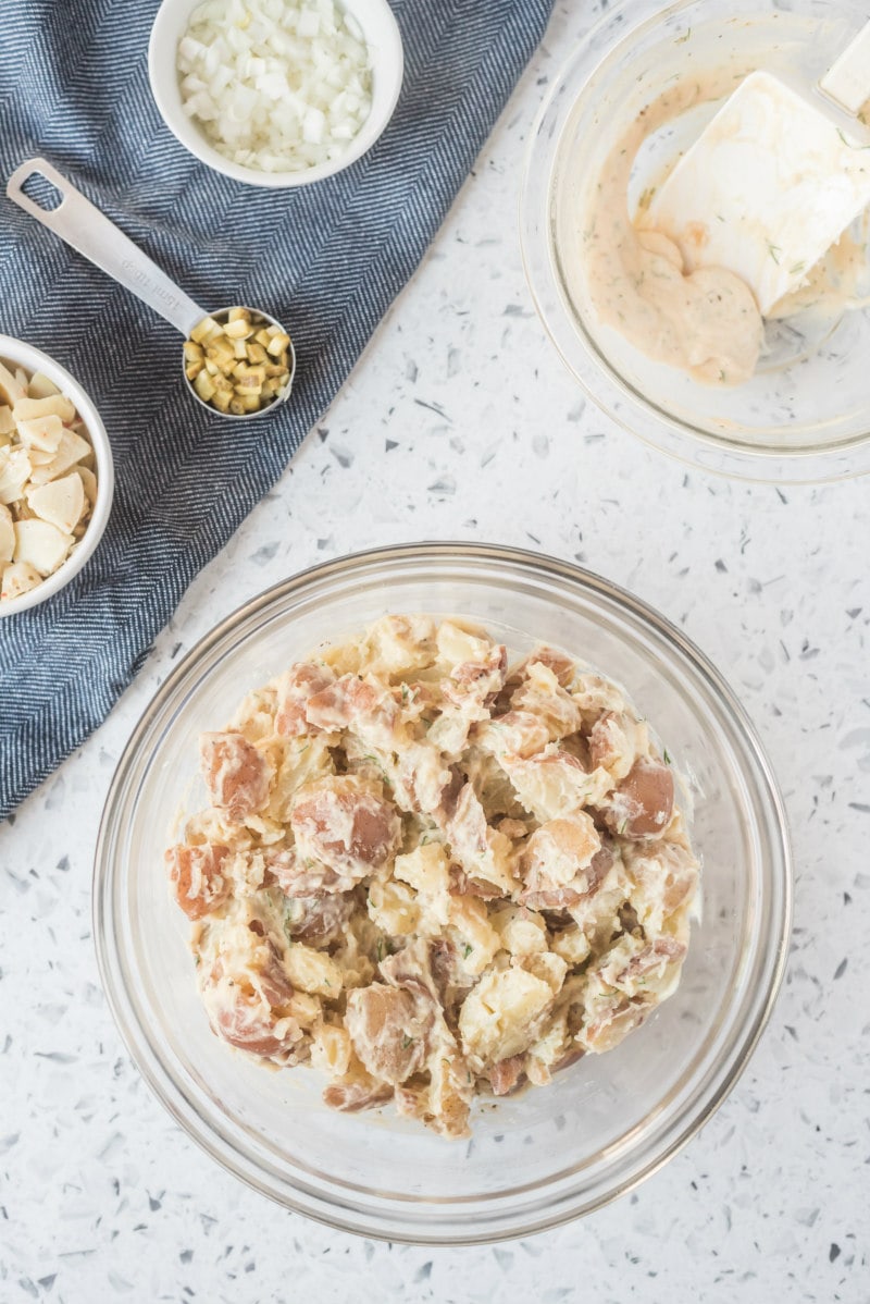 overhead shot of process of making california potato salad with potatoes mixed with dressing in a glass bowl, a few more ingredients displayed in the background in little bowls with a blue napkin