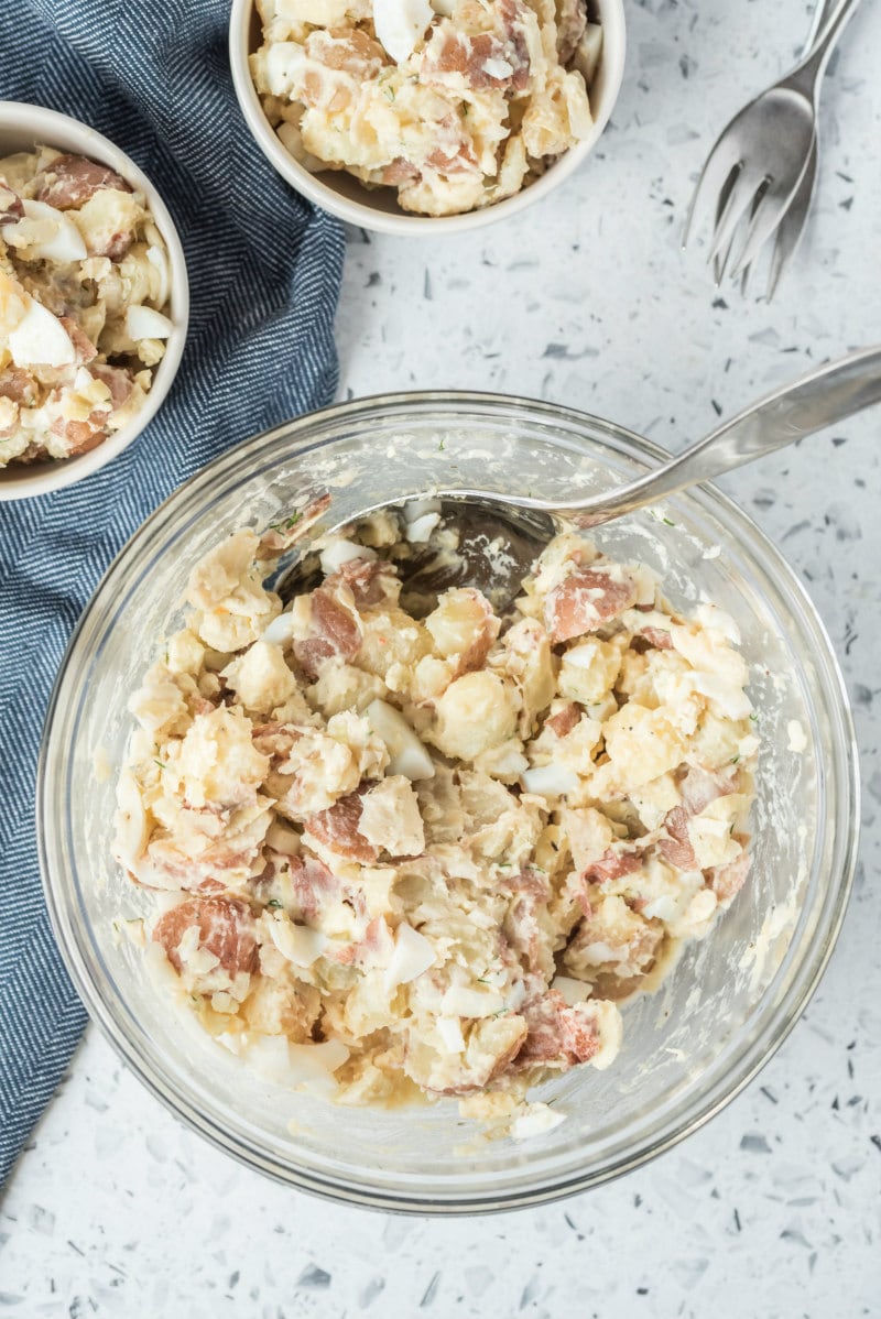 overhead shot of potato salad in a glass bowl with two servings of potato salad in white bowls on the side, set on a blue napkin. Fork in background.