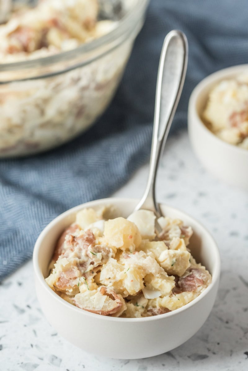 serving of potato salad in a white bowl with a spoon, full bowl of potato salad in the background set on a blue napkin