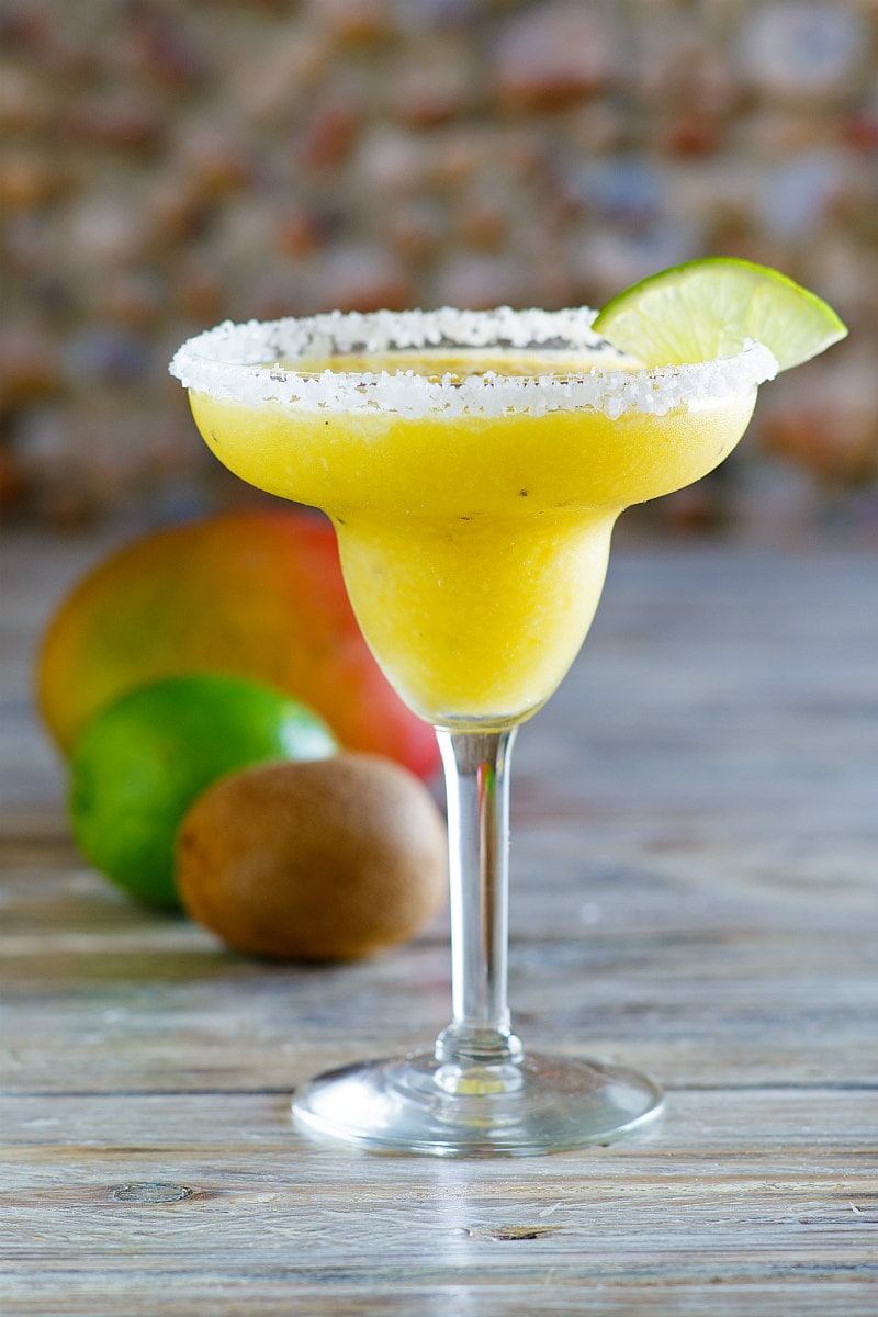 tropical margarita in a margarita glass with lime wedge and fresh fruit sitting behind