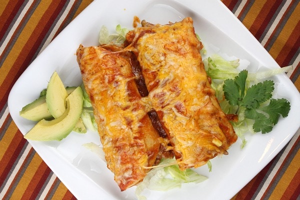 two beef enchiladas on a white plate with avocado and cilantro garnish- set on a striped placemat