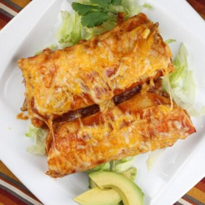 two beef enchiladas on a white plate with avocado and cilantro garnish- set on a striped placemat