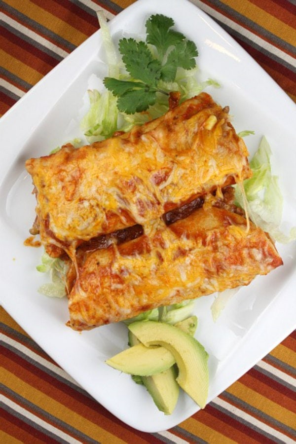 two beef enchiladas on a white plate with avocado and cilantro garnish- set on a striped placemat