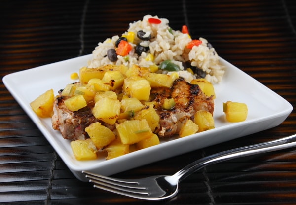 Chili Rubbed Pork Chop topped with pineapple and a side of rice on a white plate with a fork on the side