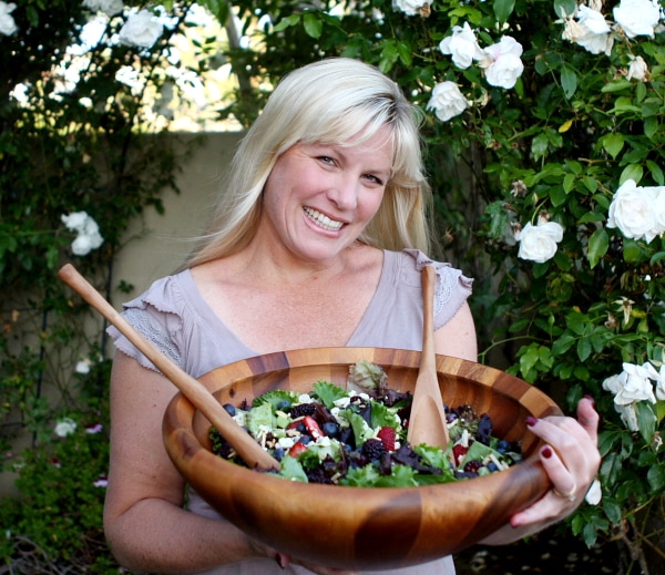 RecipeGirl holding triple berry salad in a large wooden bowl with wooden serving spoons in a backyard setting