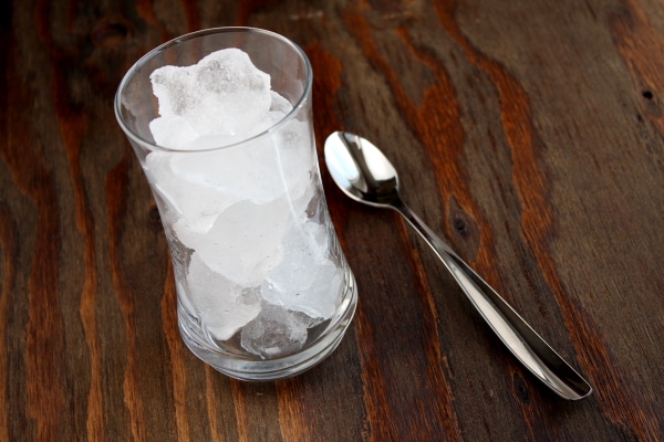 Tall glass of ice sitting on a wooden board with a spoon alongside