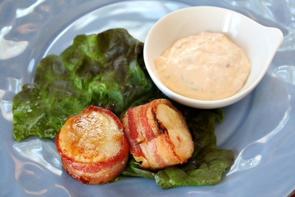 two bacon wrapped scallops sitting on a lettuce leaf on a blue plate served with a small white dish of sauce