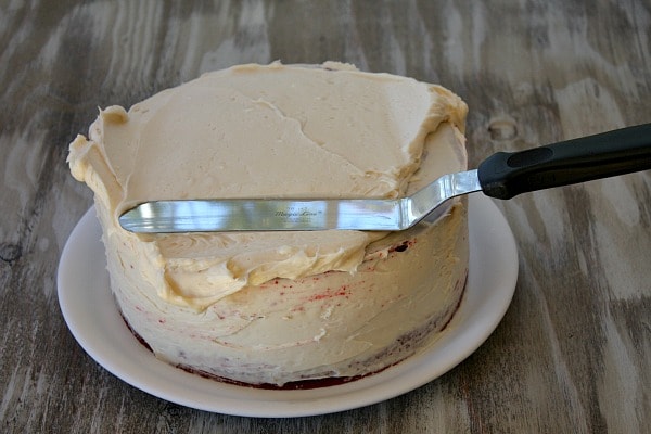 adding white frosting to a red velvet cake with a long spatula