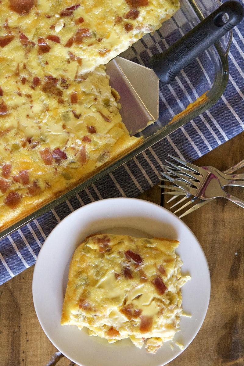 Egg Bake in a pyrex casserole dish on a blue and white striped napkin. Spatula is serving the eggs. single serving on a white plate on the side. Forks in view too.