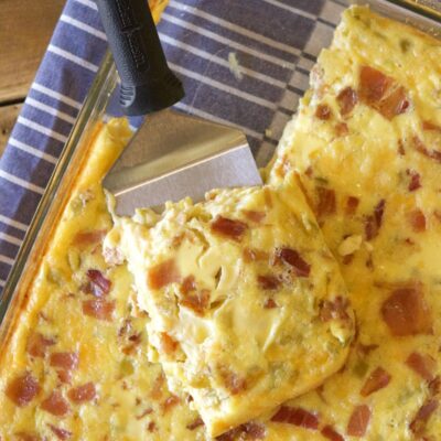 green chile bacon and cheese egg bake in a pyrex casserole dish with a spatula serving it. Sitting on a blue and white striped cloth napkin