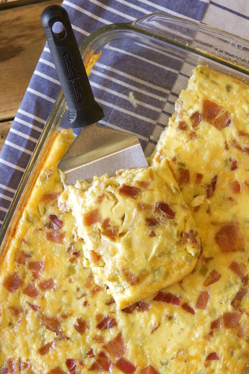 green chile bacon and cheese egg bake in a pyrex casserole dish with a spatula serving it. Sitting on a blue and white striped cloth napkin