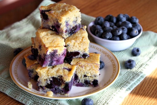 stack of slices of blueberry cornbread on a plate set on a green napkin. bowl of blueberries in white bowl on the side