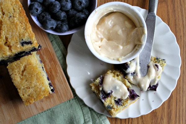 overhead shot of blueberry cornbread on a cutting board with a bowl of fresh blueberries and a slice of cornbread on a white plate with butter on it and a bowl of butter and a knife