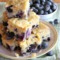 pieces of blueberry cornbread stacked on a plate with a bowl of blueberries in the background- set on a green towel on a wooden surface