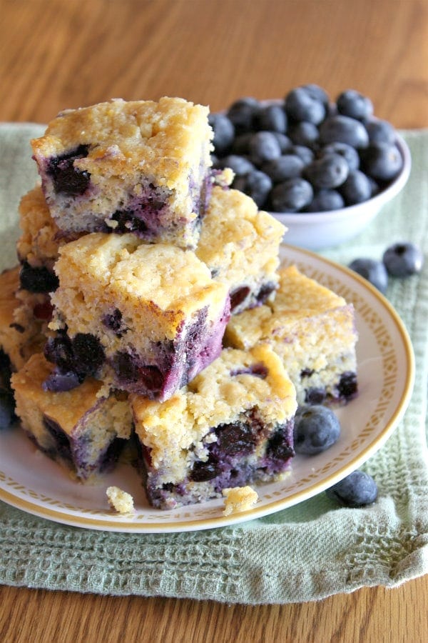 pieces of blueberry cornbread stacked on a plate with a bowl of blueberries in the background- set on a green towel on a wooden surface