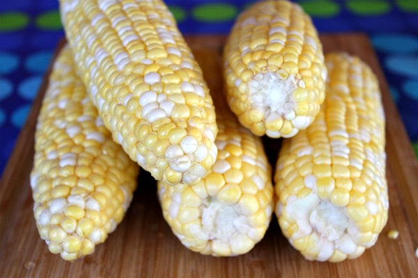 5 ears of fresh corn stacked on a cutting board with a blue and green polka dot tablecloth underneath