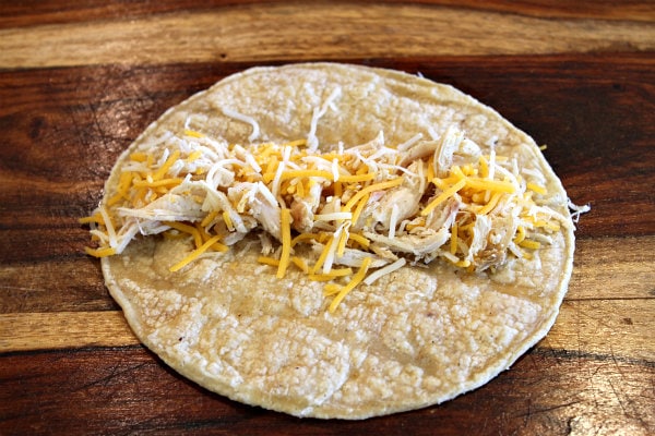 Easy Baked Chicken Taquitos recipe from RecipeGirl.com #easy #baked #chicken #tacos #taquitos #recipe #RecipeGirl How to Make Baked Chicken Taquitos showing tortilla with chicken and cheese sprinkled on top set on top of a wooden board