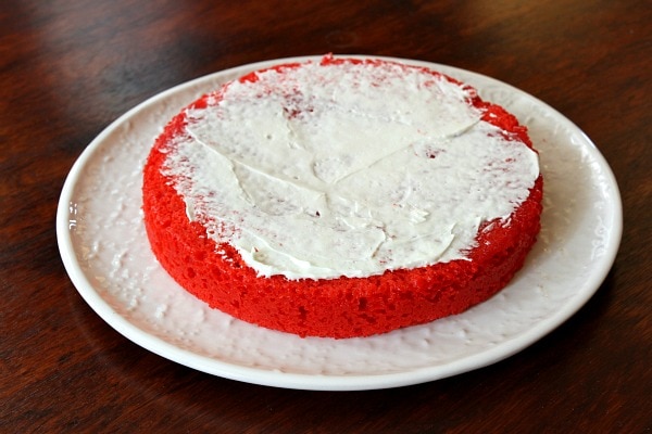 round layer of red cake on a white plate with white frosting spread on top