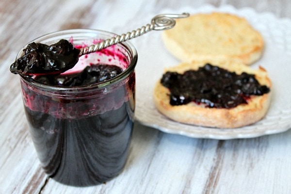 Blueberry Refrigerator Jam in a jar with a spoonful of jam sitting on top. English muffin with jam on a plate next to it.